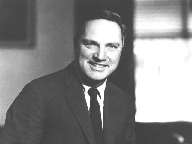 A white middle-aged man with short dark hair smiles while wearing a suit and tie.