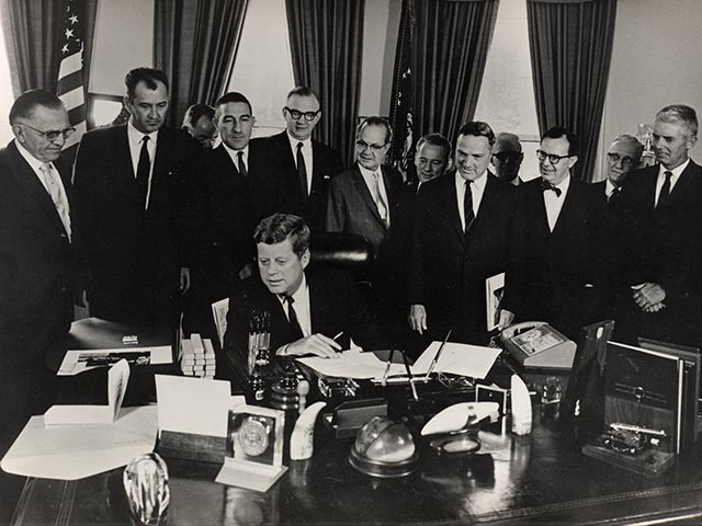 President Kennedy sits at a large desk with a pen in hand and papers in front of him. He is surrounded by 13 men in dark suits and ties.