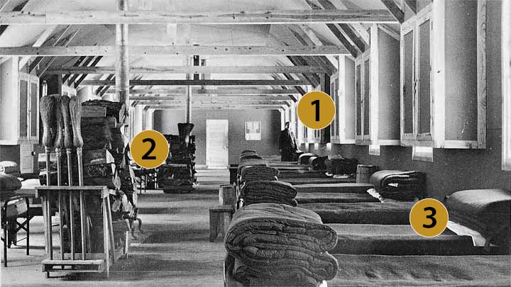 Beds lined against the wall of a barracks building with a stove in the center of the room.