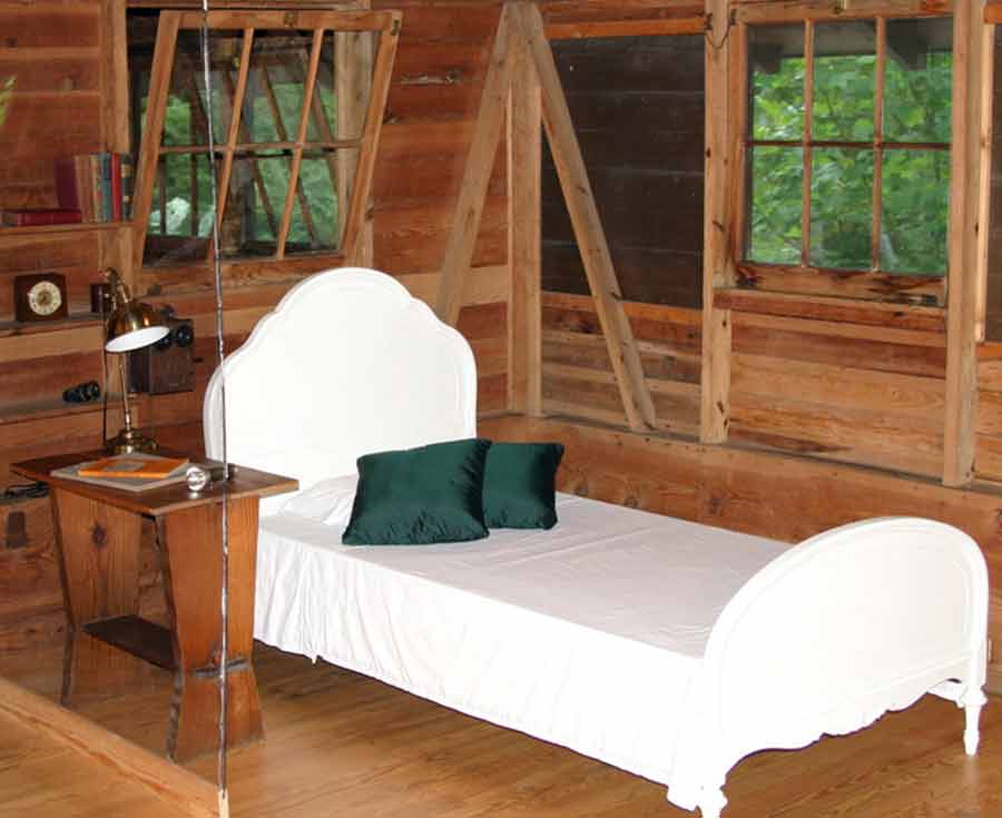 Modern day photo of the bedroom, showing brown wooden walls and floors and green pillows on the bed.
