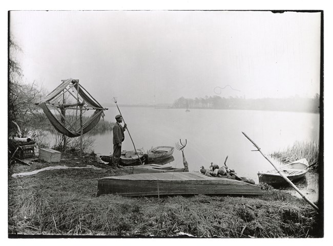 An archival black and white image featuring a gentleman with dark skin wearing a suit and holding an eel spear near a body of water with boats, decoys and other fishing equipment.