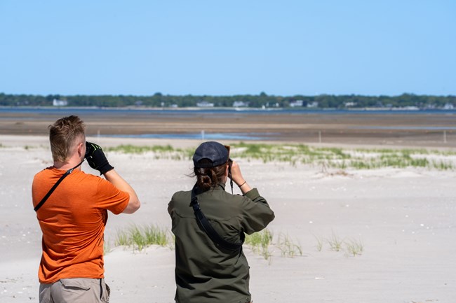 A park ranger and intern look through binoculars toward a low tide flat.