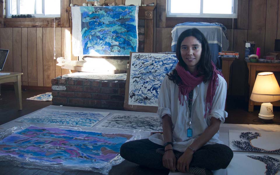 Woman with dark hair sitting on her bed with artwork spread around the room and bed in a well-lit room.