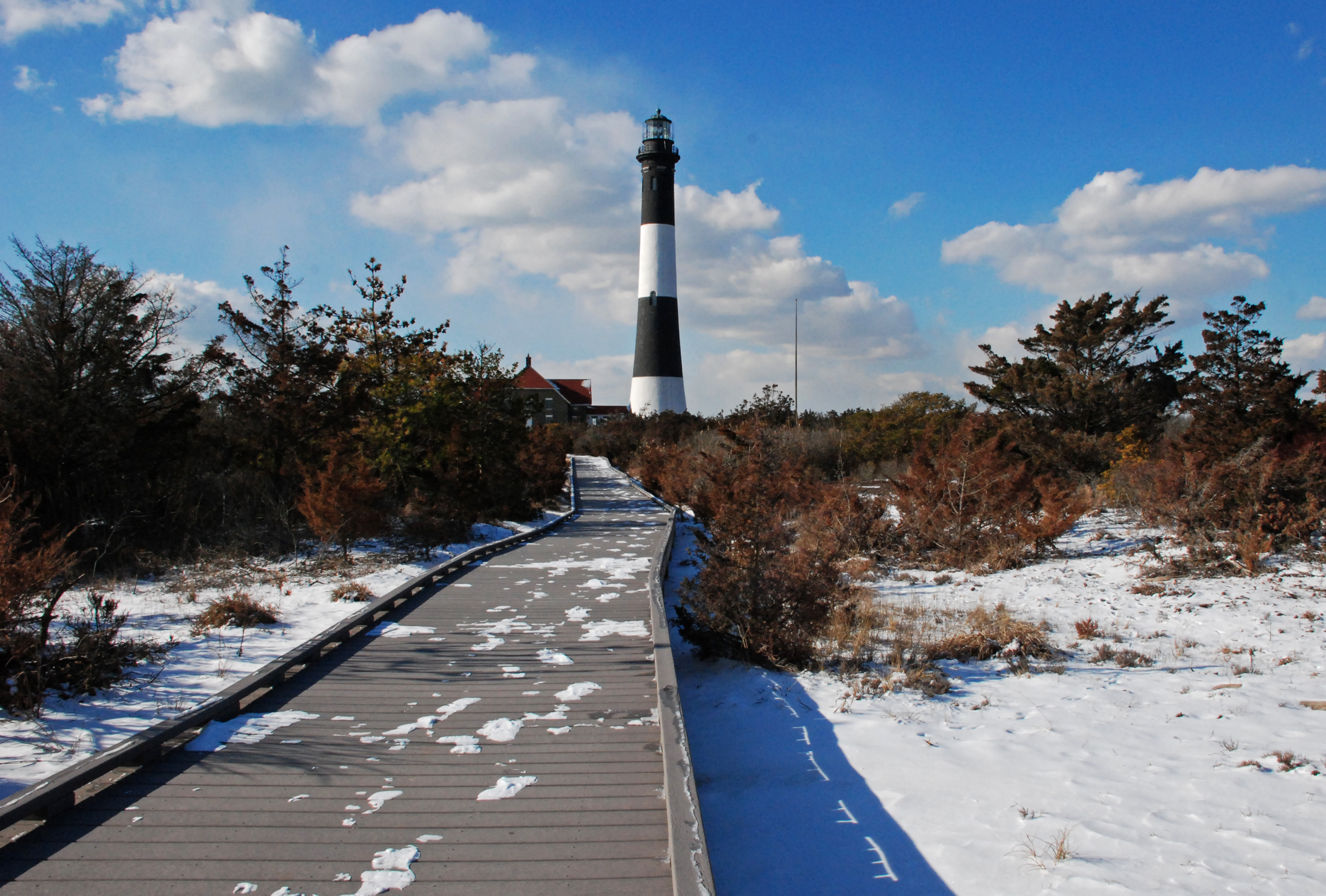 A wintry walk to the Fire Island Lighthouse. A snowy boardwalk leads to a black and white lighthouse tower.