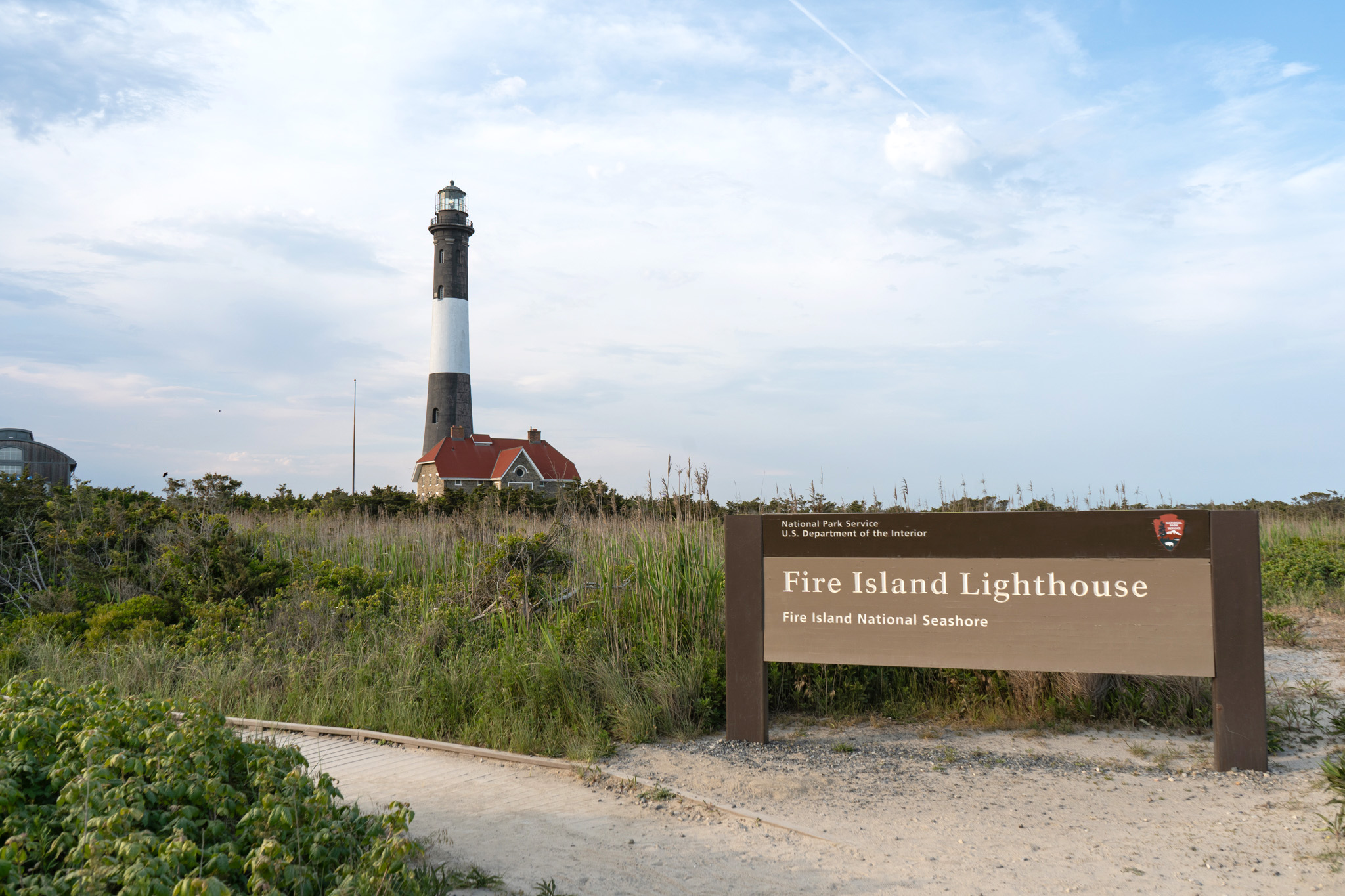 The black and white striped Fire Island Lighthouse rises in the distance. In the foregrounds is the end of boardwalk lined by vegetation and a sign reading, "Fire Island Lighthouse."