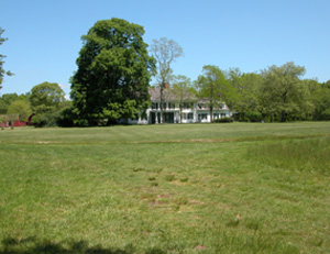 Expanse of green lawn in front of the William Floyd Estate manor house, flanked by tall leafy trees.