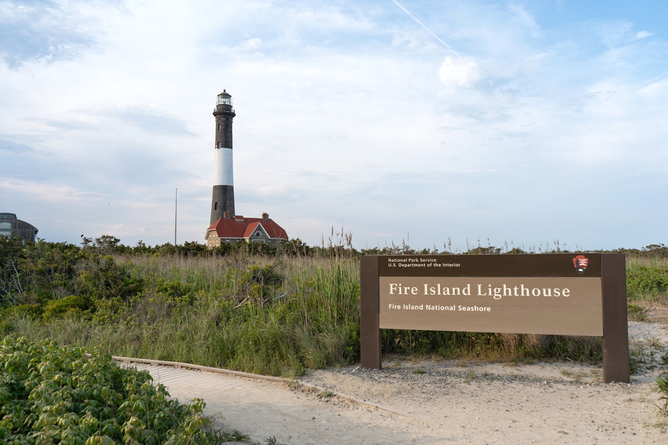 A black and white striped lighthouse in the background with a sign reading "Fire Island Lighthouse, Fire Island National Seashore," a boardwalk, and green foliage in the foreground.