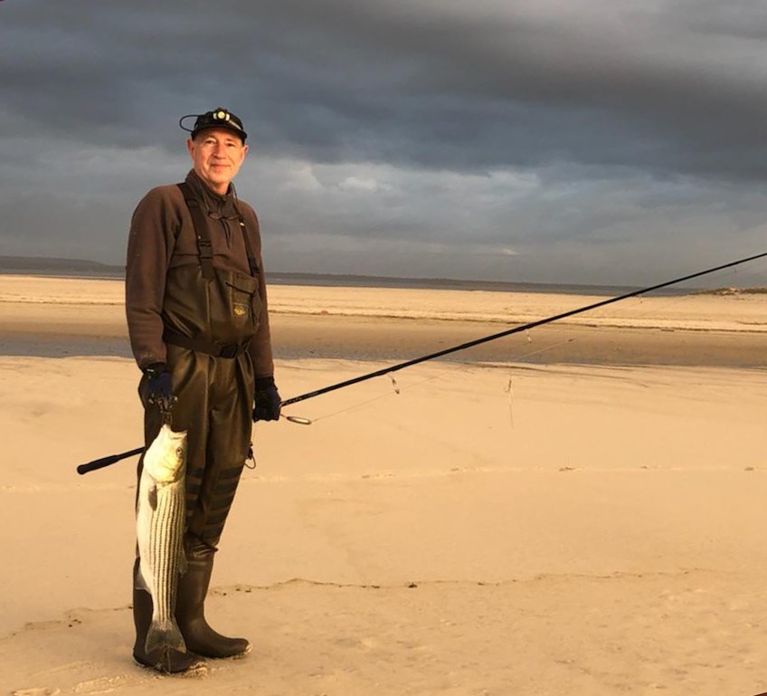 A man in waders holds a fishing pole and stripped bass near the breach.