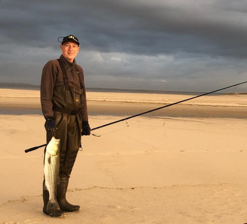 A man in waders holds a fishing pole and stripped bass near the breach.