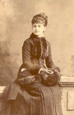 full body portrait of Mary McKee wearing furs and sitting and looking to the left