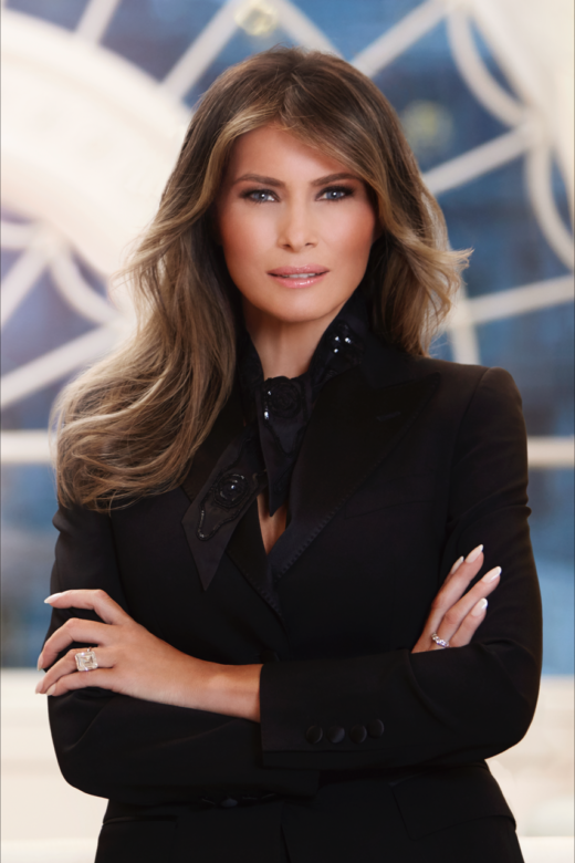 the first lady wears a black jacket and scarf and is standing with her arms crossed
