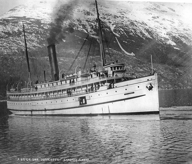 A black and white image of a steam ship with a mountain in the background.