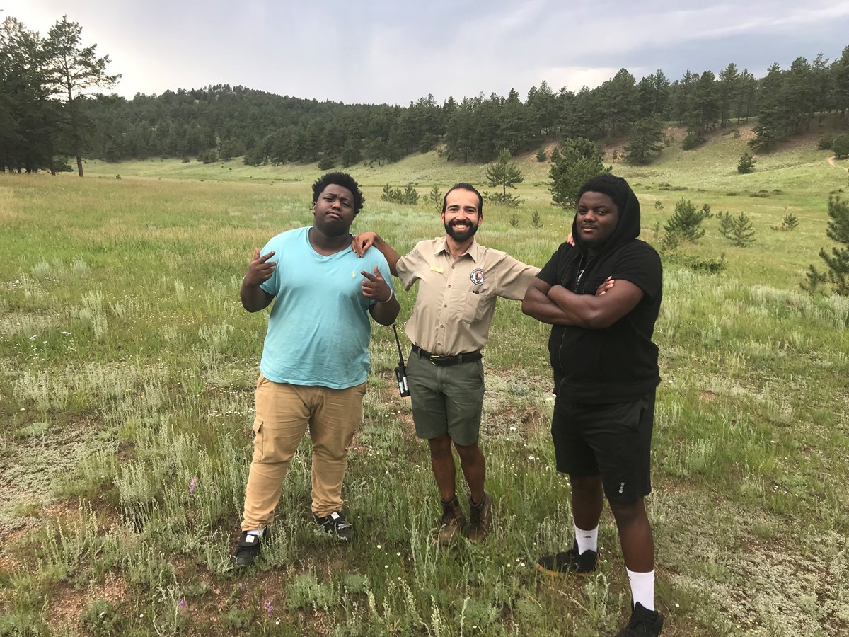 FIGURE-1-FLFO-ARTICLE-PARK-PALEO-NEWSLETTER Education Intern, Kevin Jauregui stands next to hip hop artists Nehemiah Vaughn and Joey Lovett in a grassy meadow at Florissant Fossil Beds.
