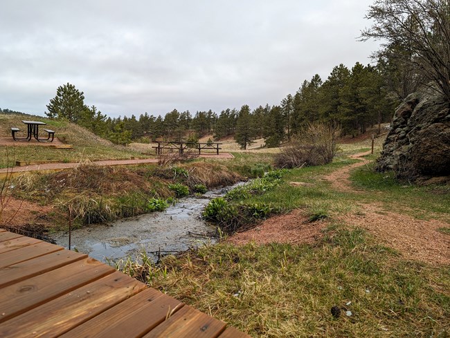 A bridge leads over a stream to several picknick tables. A trail winds past a rock formation on the right toward a hillside covered in pine trees.