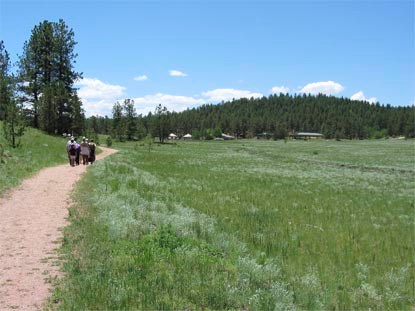 People hiking on a gravel hiking trail past a green grassy meadow with pine covered hills in the background. People hiking on a gravel hiking trail past a green grassy meadow with pine covered hills in the background.