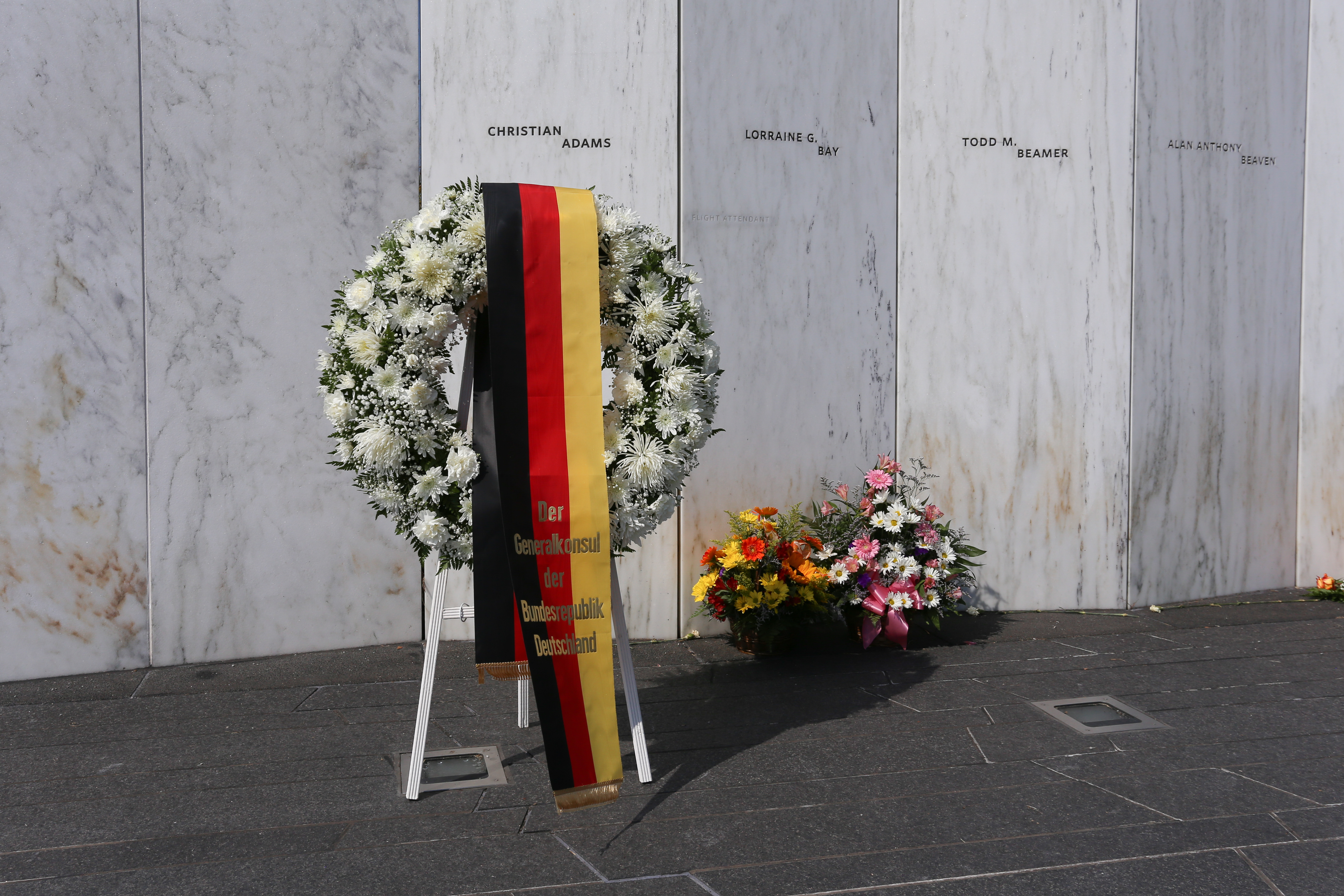 A white wreath draped with German Flag and text sits in front of a white marble wall inscribed with the names of the passengers and crew member of Flight 93