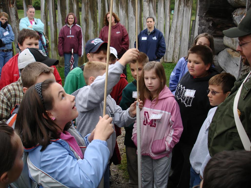 Kids, parent, and a park ranger in a wooden fort placing wooden dowels into white plastic tubes to replicate loading a musket