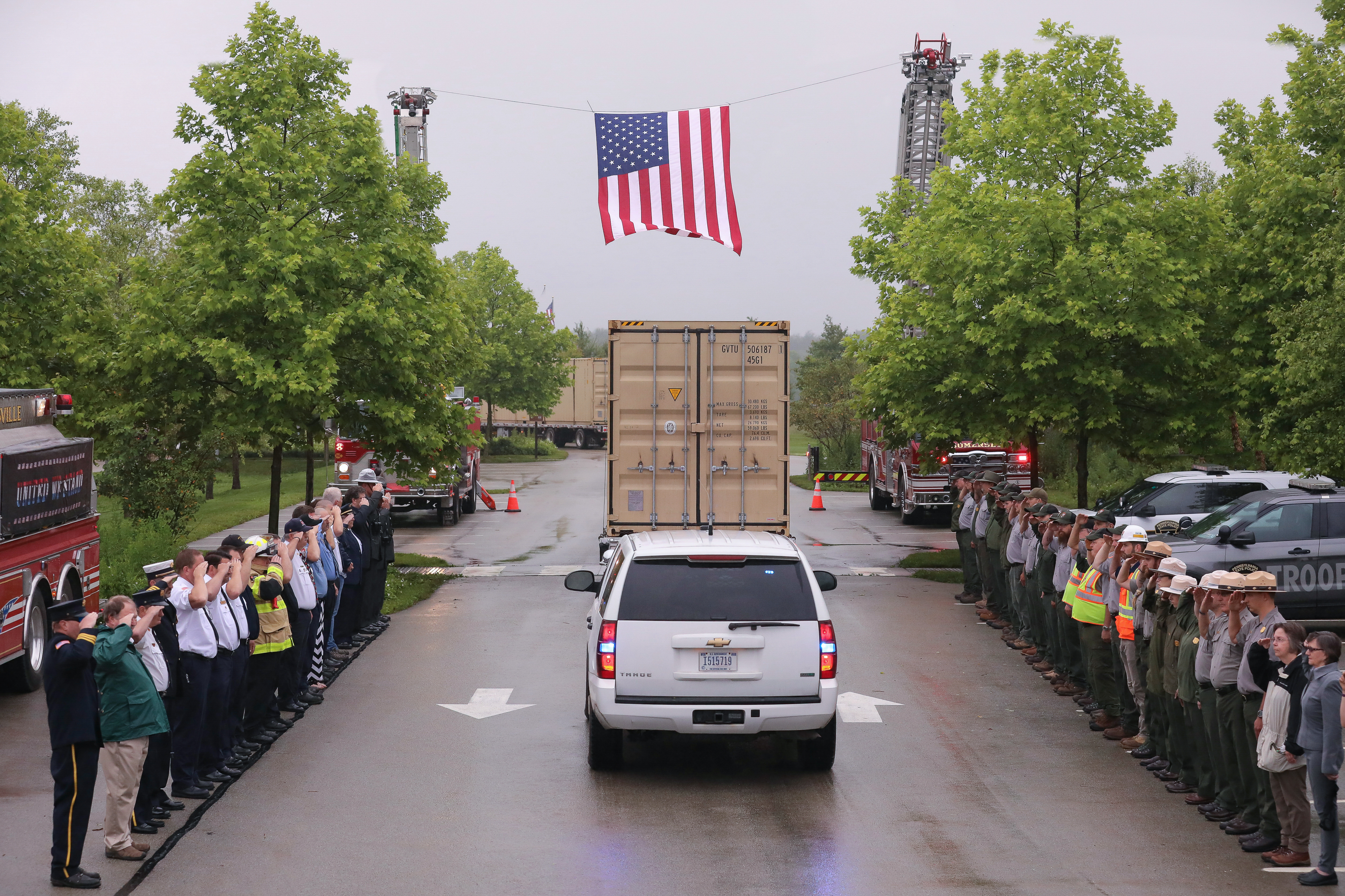 Flatbed truck with a large shipping container drives under a suspended American flag as onlookers salute. Flatbed truck with a large shipping container drives under a suspended American flag as onlookers salute.
