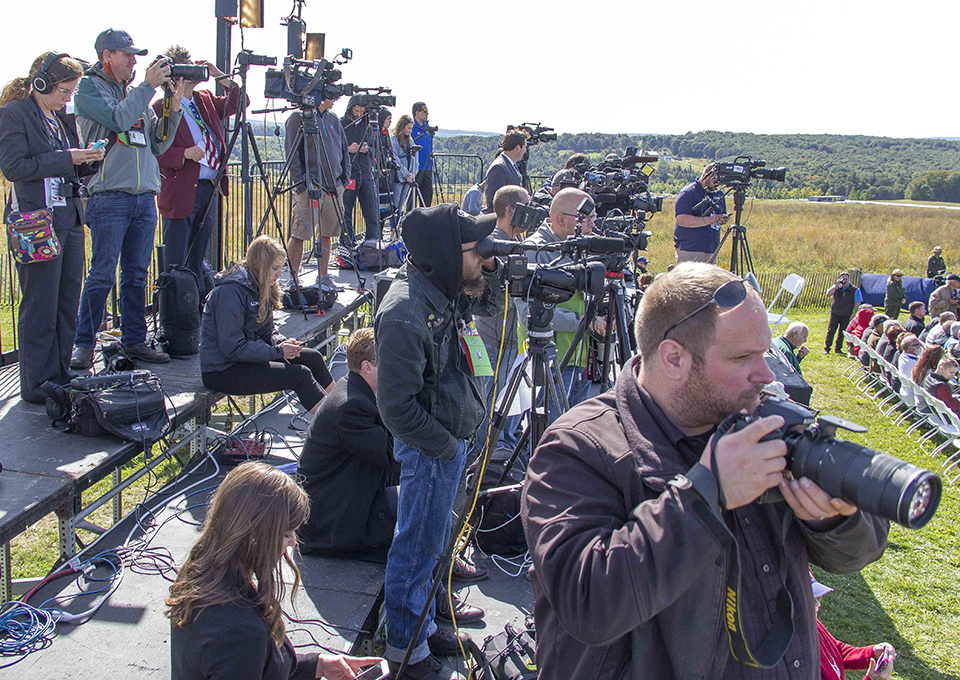 Members of the press stand on a riser at the back of the crowd during an anniversary event. Members of the press stand on a riser at the back of the crowd during an anniversary event.