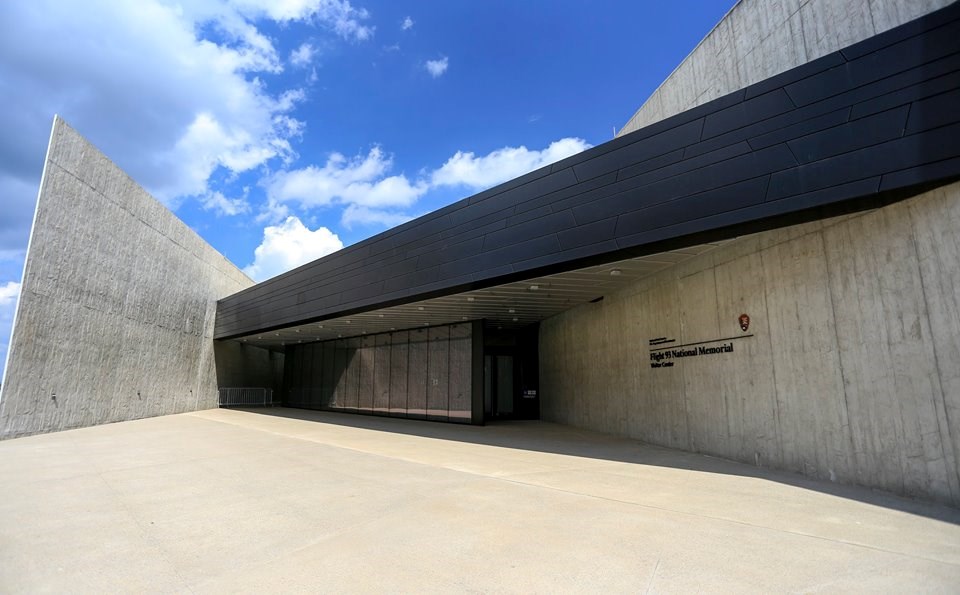 Blue skies appear over the entrance of the visitor center with glass windows and grey concrete walls stamped to appear like hand hewn logs.