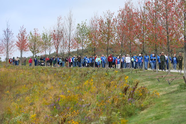 People walking on a trail next to field of wildflowers and trees in autumn with red and orange leaves..
