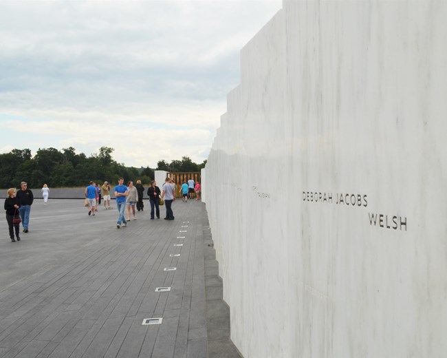 Visitors walking to white marble wall, black granite walking surface marking linear flight path and wooden hand cut gate.