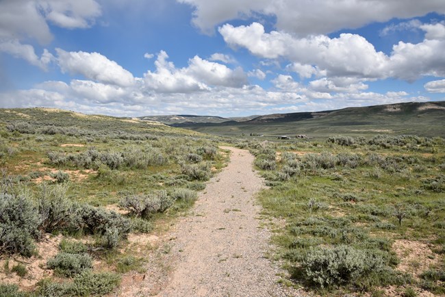 A dirt path through sagebrush leads straight away from the viewer with small buildings visible in the distance on the right. The sky is bright blue with puffy white clouds.