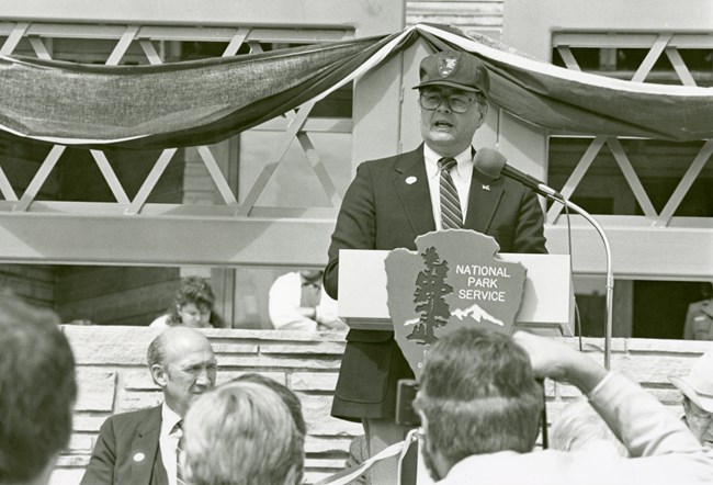 A man in a suit and an NPS ballcap stands at a podium with the NPS arrowhead. There is a few people visible in front of him watching and bunting hanging from the building behind.