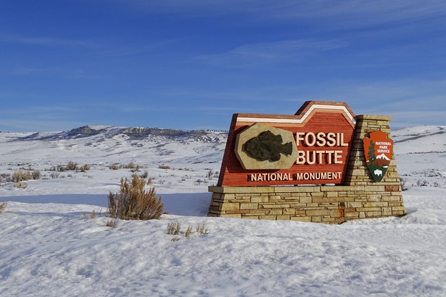 A sign that says Fossil Butte National Monument with an image of a fish fossil and the NPS arrowhead.  Snow and some sagebrush surround the sign and there is a bright blue sky above.