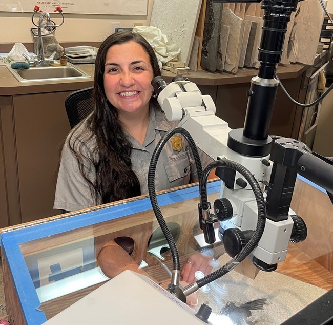 A park ranger smiles at the camera. She sits at a microscope with a fossil underneath.