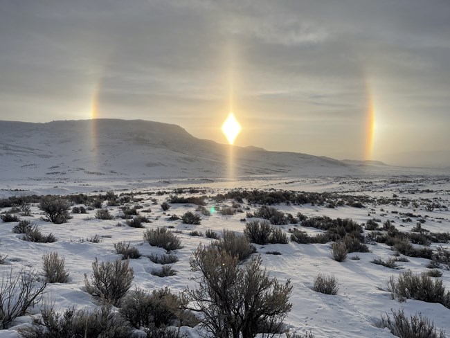 A snowy landscape with the butte towards the left. The sun is in the middle with large rays going up and down and a quarter of the photo away on each side are rainbow semi-circles arcing towards the sun.