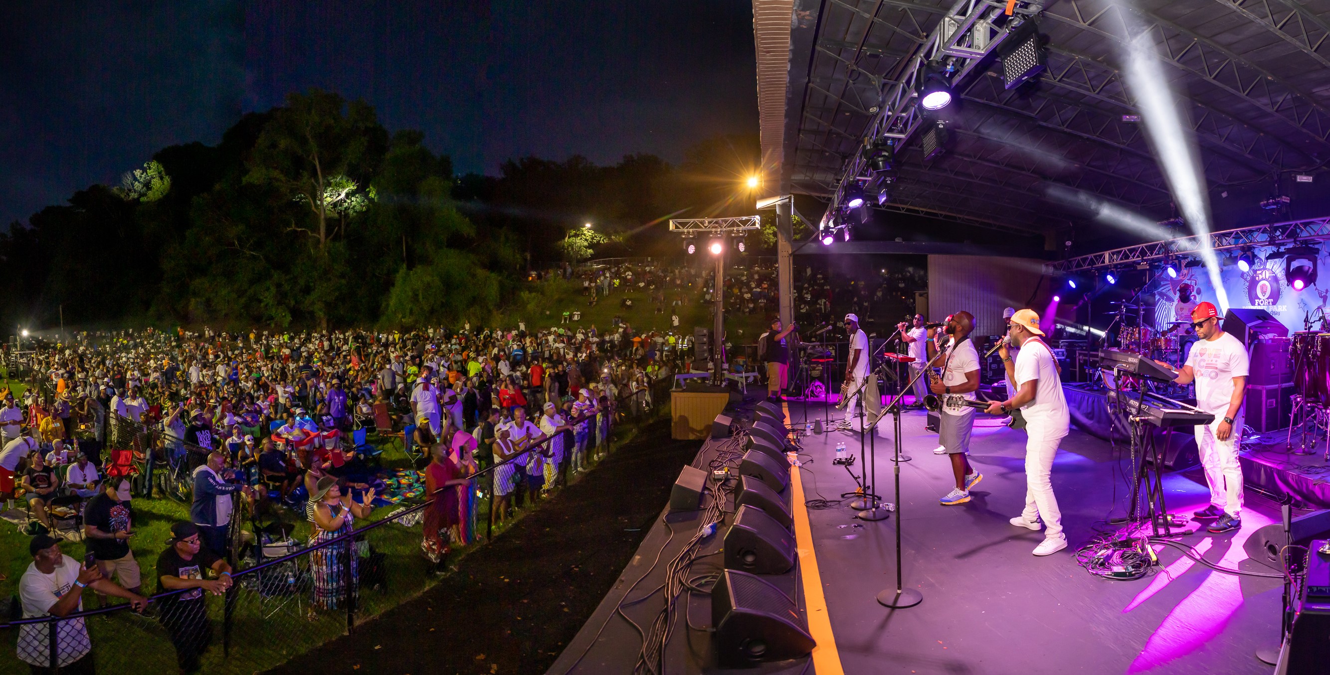 Fort Dupont Amphitheater A band wearing white shirts bathed in purple light performs on stage for a crowd on a lawn at night.