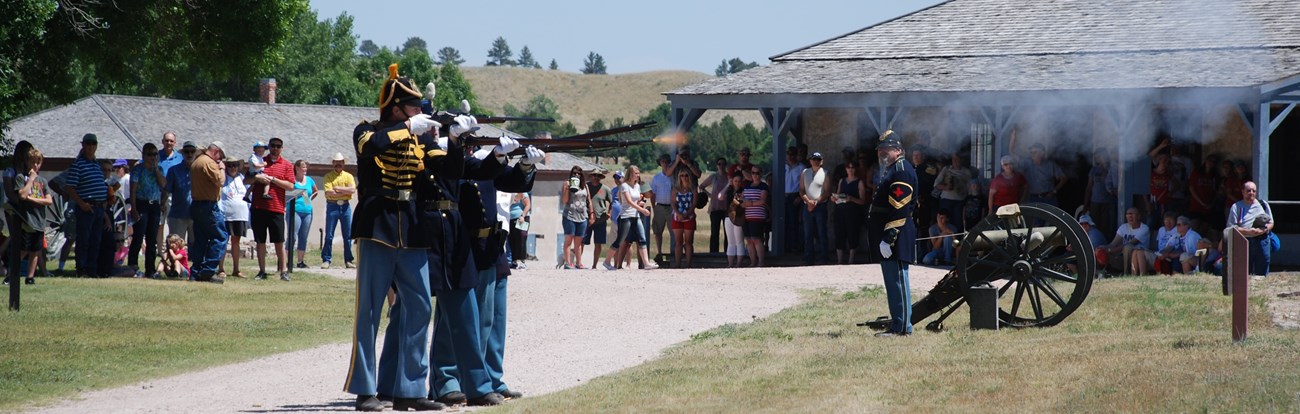 People in historic army uniforms fire rifles while visitors and an enlisted artillery soldier watch.