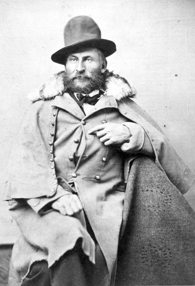 A man in a fur coat and hat is seated on a chair, presenting a sophisticated and elegant look.