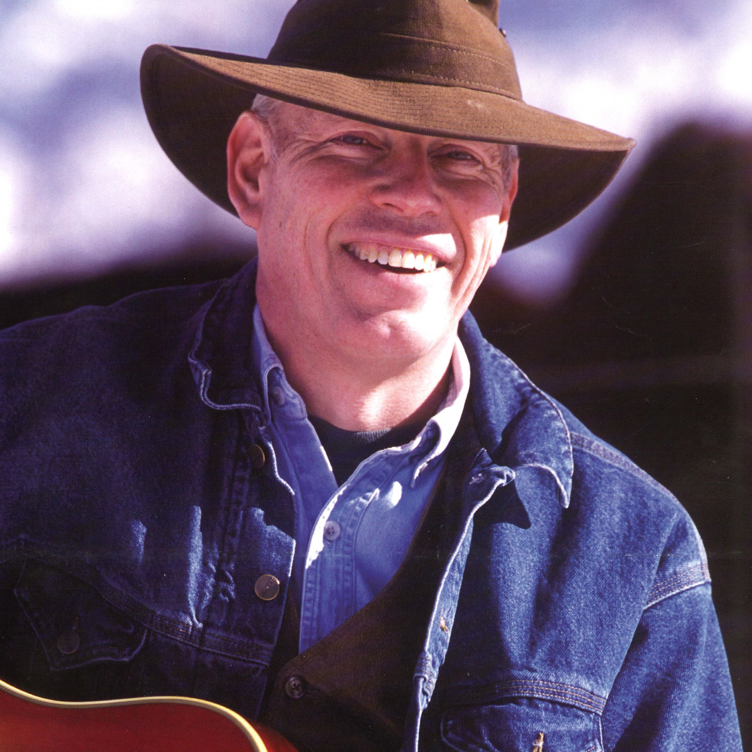 2022 Hank Cramer Poster-CROP A smiling man wearing a broad-brimmed hat and holding a guitar.