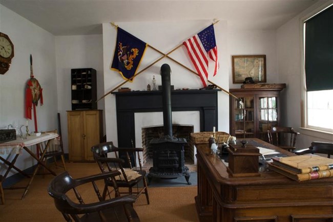 Inside the officer quarters room. The room includes a large brown desk, four chiars, several shelves, and a fireplace with two falgs on the mantel.