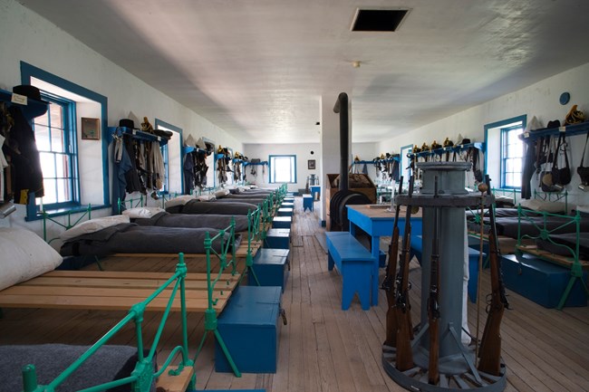 The inside of the Calvary Barracks. The room contains beds lined up agaisnt eachother, equiment haning on the walls, and miltary guns in the middle.