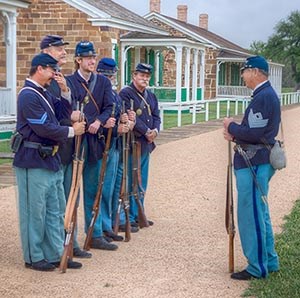 Men in 19th century U.S. Army uniforms lined up in front of sandstone quarters. Men in 19th century U.S. Army uniforms lined up in front of sandstone quarters.