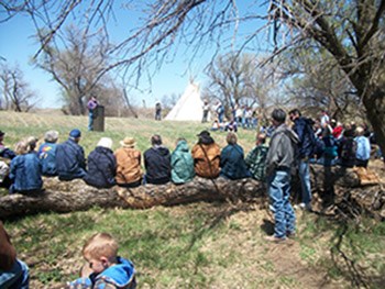Group of people listening to a talk outside with a teepee in the background.