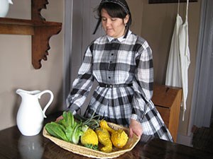 Woman in 19th century dress hold basket with lettuce and squash.