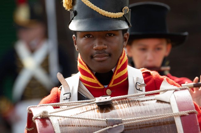 Drummer in red War of 1812 uniform looking over bass drum.