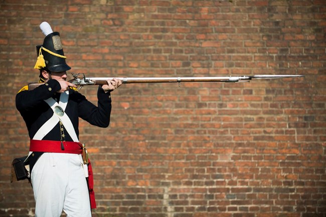 Soldier in blue uniform aiming musket. Fort in background.