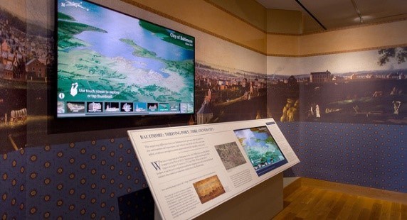 Photograph of an interior exhibit showing a monitor on a wall and an exhibit panel.