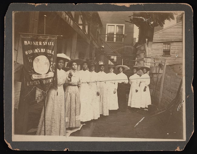 A black and white photo of a group of African American women holding a banner that reads "banner state woman's national Baptist Convention"