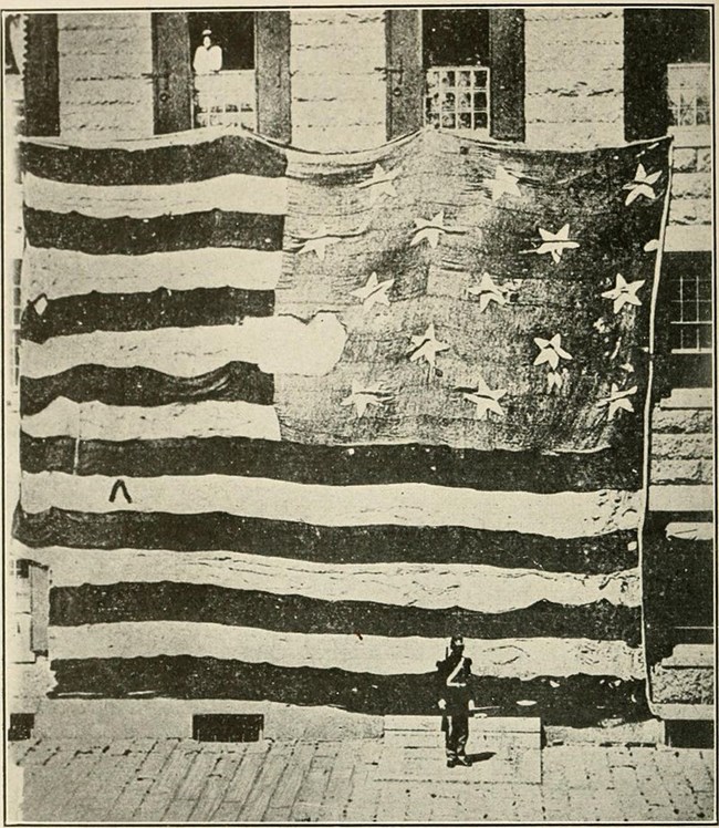 A historic black and white photograph of the large Star-Spangled Banner flag on display outside of a building with a soldier standing in front of it.