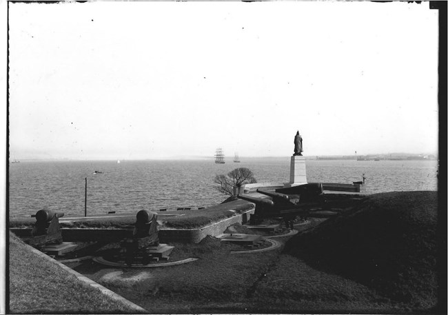 A black and white photograph looking from the fort towards the Potapsco river with cannons and the George Armistead statue in the foreground.