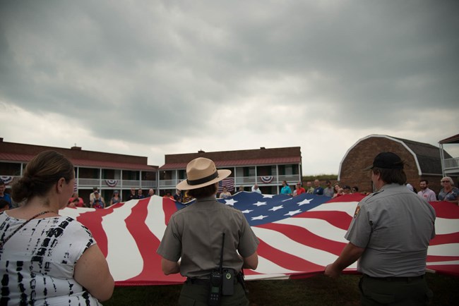 A ranger talking to a group of visitors all holding on to a large 15 star, 15 stripe American flag inside the star fort.