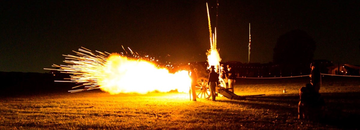 a group of living historians fire a cannon at night with the fort in the background.