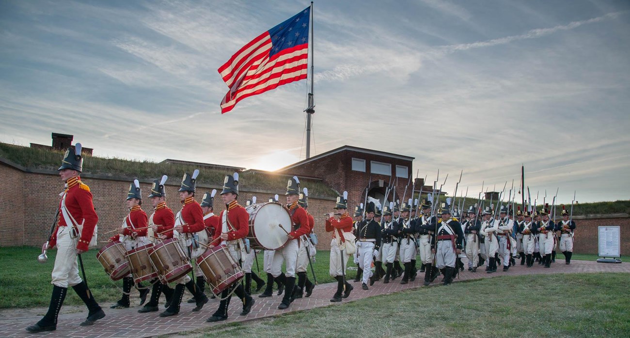 Musicians and soldiers in War of 1812 uniform march in front of brick fort with 15-star American flag flying above.