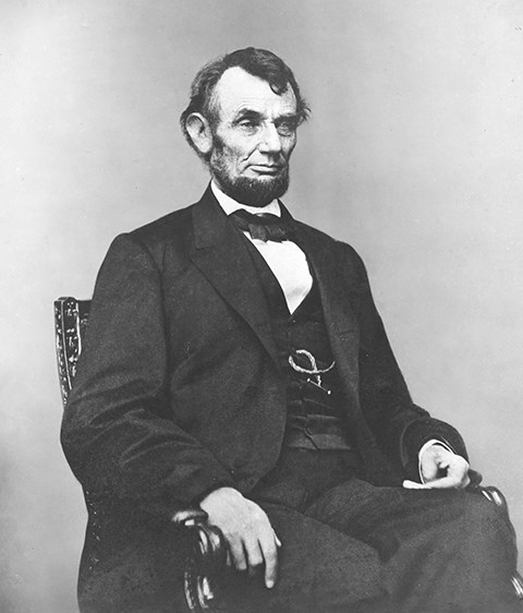 Abraham Lincoln poses for a picture. He is sitting in a chair wearing a black suit.